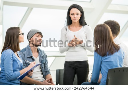 Young woman is sharing her problems with people. View of woman is telling something and gesturing while group of people are sitting in front of her and listening.