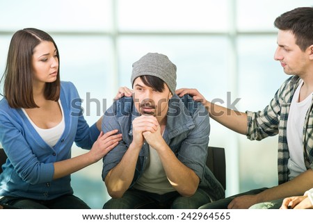 Depressed young man is sitting at the chair while two other people are comforting his.