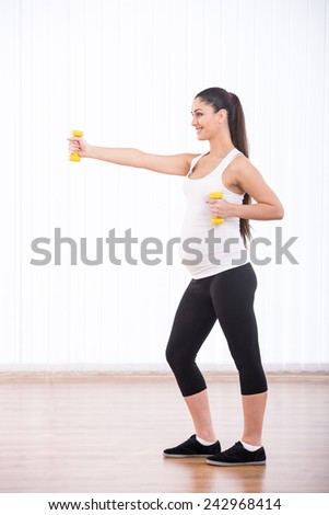 Beautiful, smiling pregnant woman is doing sport with dumbbells, isolated on white background. Side view.