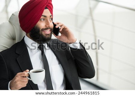 Smiling indian businessman is drinking coffee in his office and talking by phone.