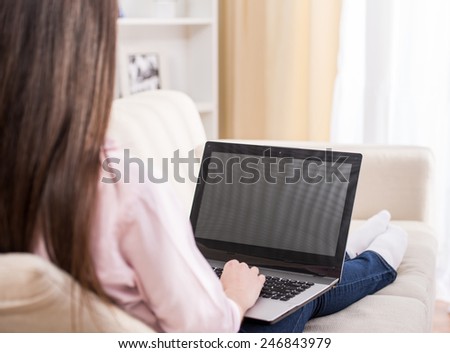 Back view of young woman is using laptop while sitting on couch at home.