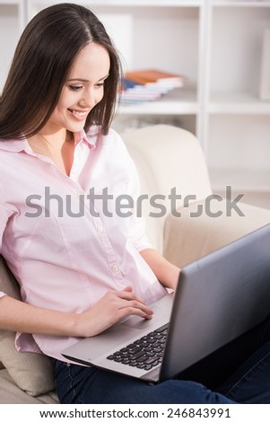 Casual young woman is using laptop while sitting on couch at home.