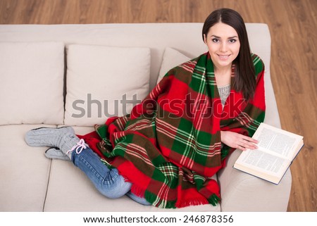 Beautiful young girl sitting on sofa at home wrapped a blanket and is reading.