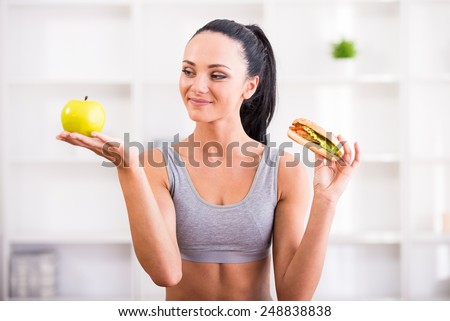 Fitness, home and diet concept. Young woman with apple and hot dog after exercising at home. Difficult choice.
