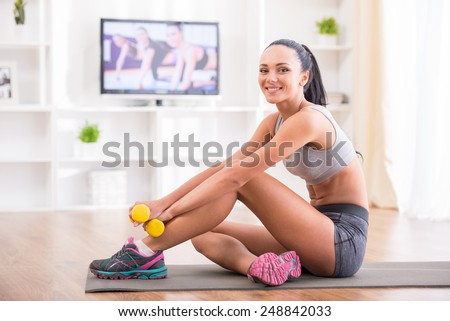 Fitness at home concept. Smiling young woman is sitting on mat with sports equipment at home.