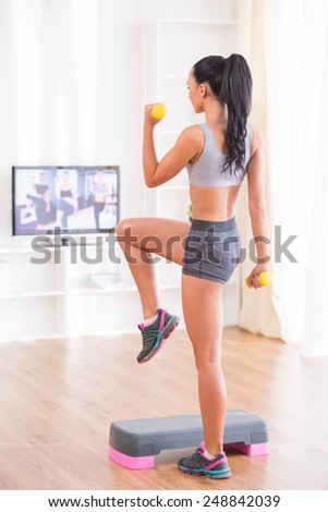 Young woman is exercising with dumbbells and step at home.