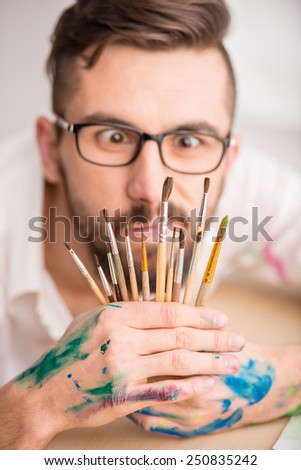 Portrait of a young male artist with many brushes for painting is looking at the camera through them.