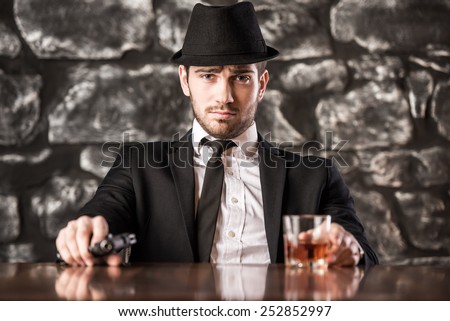 Confident, gangster man in suit and hat is sitting at the table with a glass of whisky and gun.