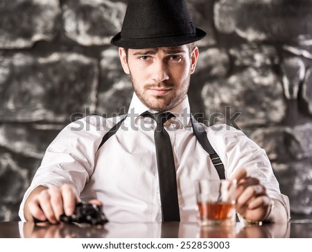 Confident, gangster man in shirt, suspenders and hat is sitting at the table with a glass of whisky and gun.