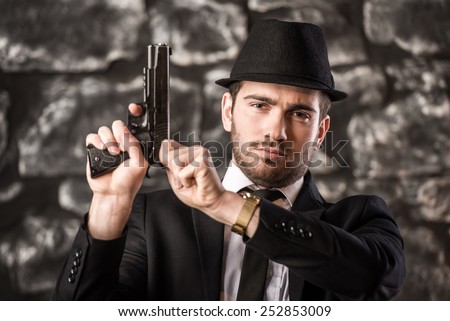 Confident, gangster man in suit and hat is sitting at the table with a gun.