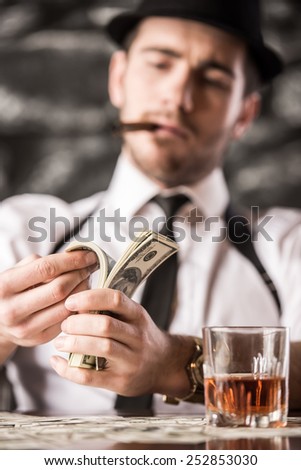Gangster in shirt and suspenders is counting money and smoking Cuban cigar, while sitting at the table.