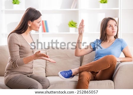 Teen girl is showing stop gesture to angry mother while sitting on sofa at home.