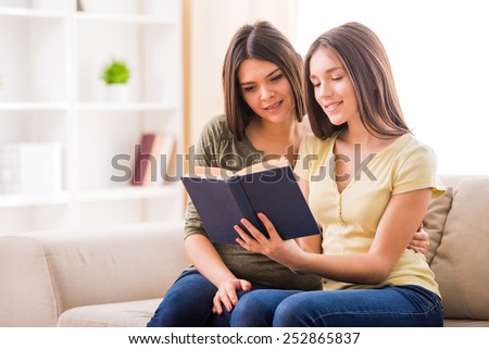 Beautiful mother and her cute teen daughter are reading a book together while sitting on sofa at home.