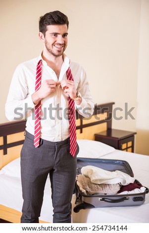 Smiling young man in white shirt is tying his necktie and looking away. Suitcase with things on the bed.