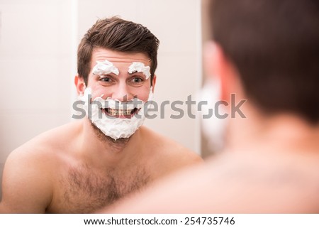 Shaving with fun. Rear view of playful young man with shaving cream on his face is standing in front of the mirror and smiling.
