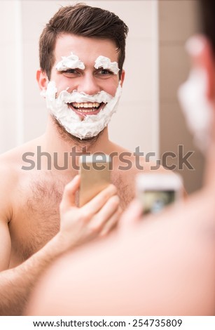 Shaving with fun. Rear view of playful young man with shaving cream on his face is standing in front of the mirror and making selfie.