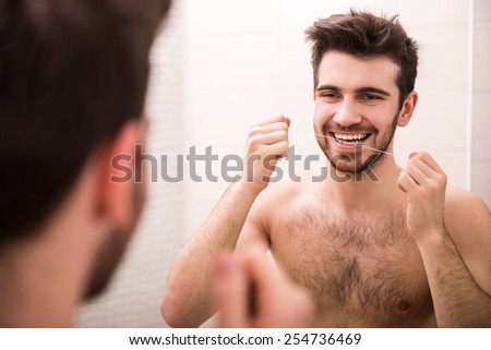 Rear view of young, smiling man is cleaning teeth with dental floss while looking at the mirror.