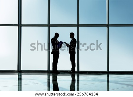 Silhouette view of two young businessmen are standing in modern office with panoramic windows.