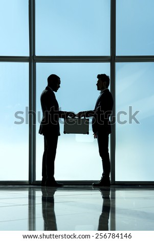 Silhouette view of two young businessmen are standing with suitcase in modern office with panoramic windows.