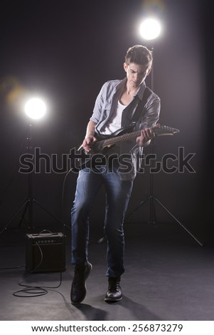 Guitar player. Young man is playing guitar against in dark room with lights behind him.