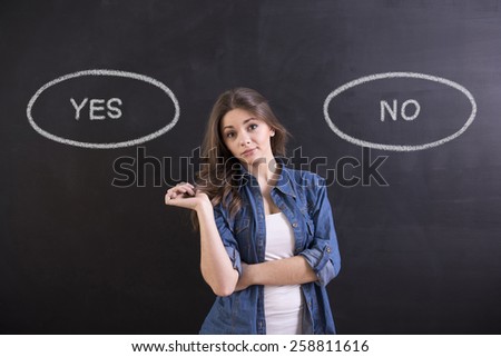 Young woman is standing on blackboard background and thinking: yes or no.