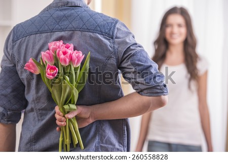 The young man is hiding flowers behind their backs to his girlfriend at home.