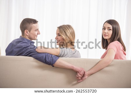 Love triangle. Beautiful young woman holding hands with man sitting near his girlfriend on sofa.