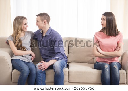 Jealousy. Young sad women sitting on the couch with her arms crossed while another women and men hugging near her.
