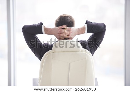 Businessman relaxing in the chair of his office with his hands behind his head.View from the backside.