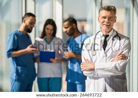 Portrait smiling mature male doctor standing with arms crossed. The background doctors speaking.