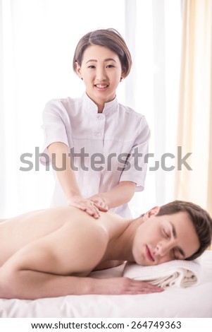 Female asian masseur doing back massage on man body and looking at camera in the spa salon.