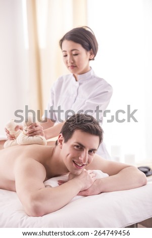 Asian female masseur doing massage on male body with spa herbal balls in the spa salon.