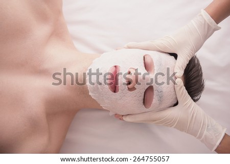 Spa therapy for young male receiving facial mask at beauty salon.