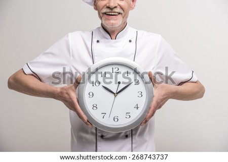 Close-up. Time to dinner. Senior male chief cook in uniform holding a clock on grey background.