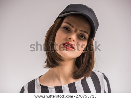 Portrait of youngn beauty woman in black-white shirt and in cap looking at camera on grey background.