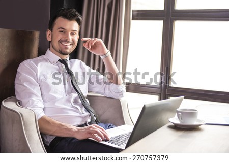 Side view. Handsome young businessman working at laptop in restaurant and looking at camera.