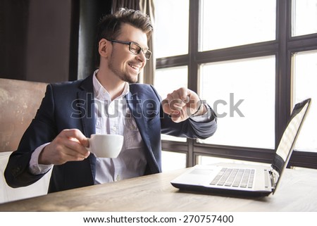 Handsome young businessman working at laptop with cup of coffee in restaurant. And looking at watch.