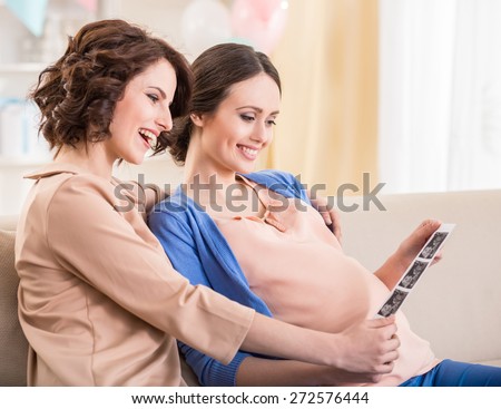 Woman is visiting pregnant friend at home. They are looking at  x-ray image of baby.