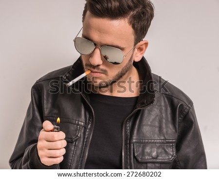 Young stylish man in glasses is smoking a cigarette. Gray background.