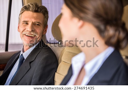 Mature handsome businessman in suit sitting in the car with his beautiful personal assistant and talking to her.