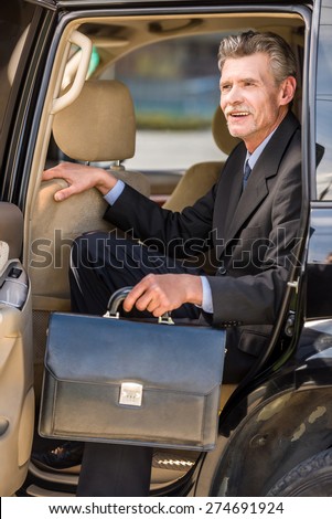 Mature smiling handsome businessman in suit  coming out of a car, holding a briefcase.