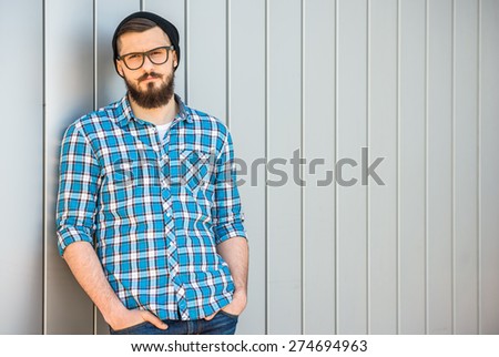 Young confident man with beard in glasses and hat is looking at the camera. Outdoor.