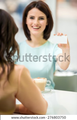 Young beautiful girl sitting in cafe with her friend and showing  new business card.