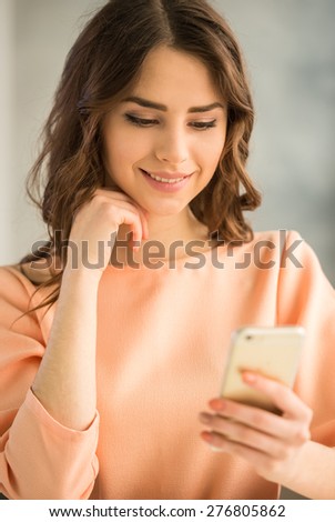 Young beautiful girl in peach blouse reading messages on smart phone on grey background.