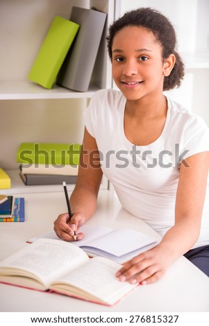 Young pretty smiling schoolgirl sitting at the table and writing homework on colorful background.
