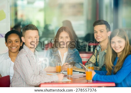 A group of teenagers sitting at the table in cafe, studying and drinking orange juice. Image through a glass.