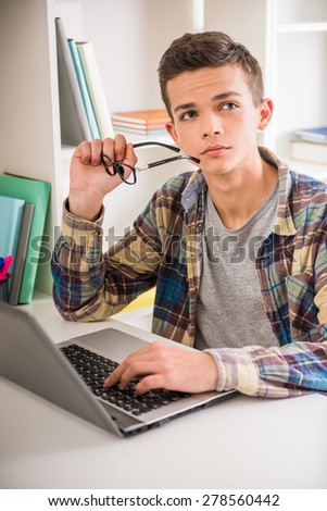 Male teenager sitting at the table at home, using laptop and thinking about something.