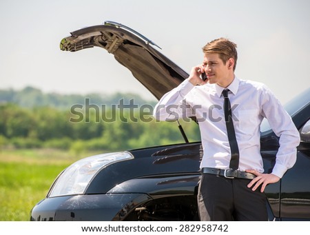Young man having trouble with his broken car, opening hood and calling for help on cell phone.