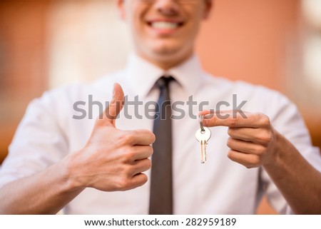 Successful realtor in white shirt showing key and making thumb up at camera.