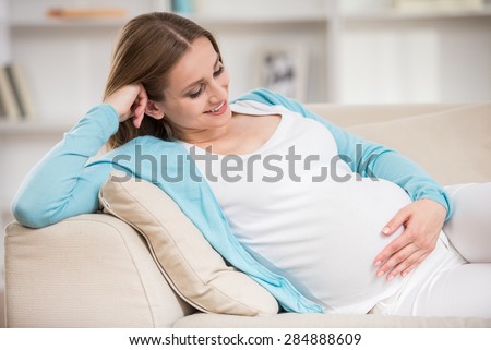 Beautiful pregnant woman sitting at sofa and keeping hand on belly.